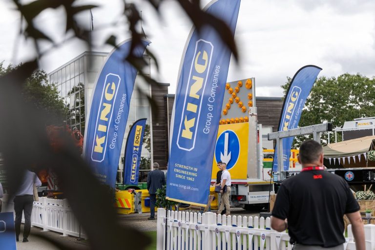 Blue KING flags flutter at an outdoor trade show, with a truck and people near a white picket fence.