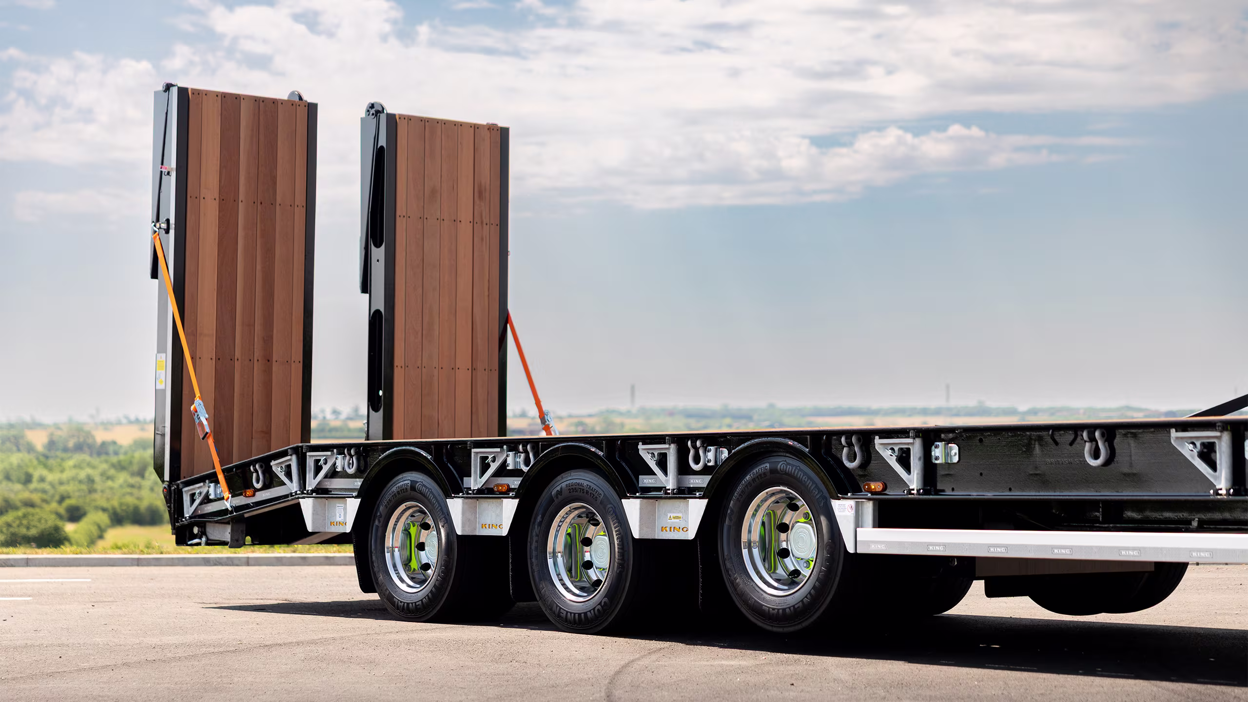 Flatbed trailer carrying tall vertical wooden panels secured with orange straps, parked on a highway with a rural landscape in the distance.
