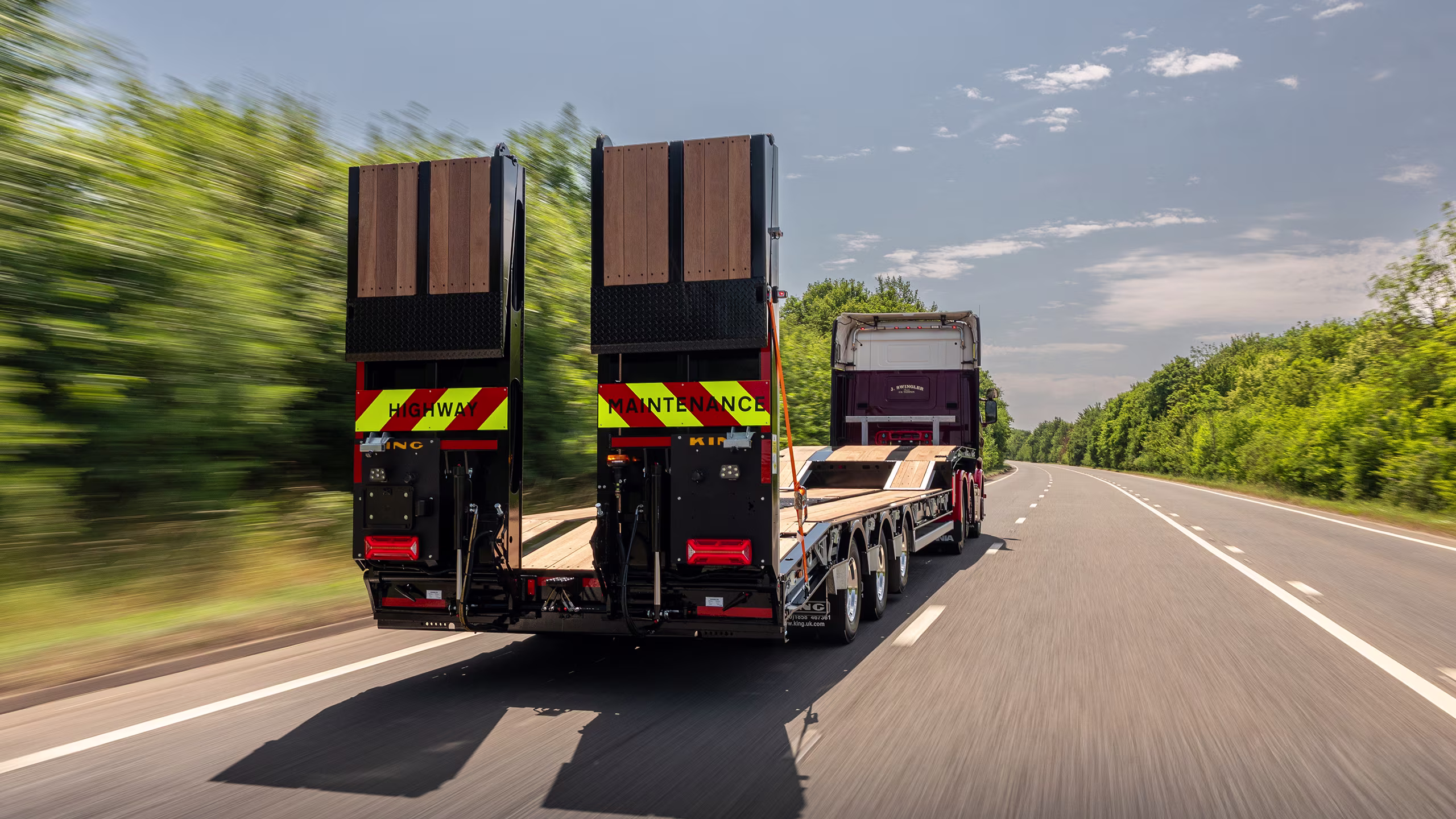 Rear view of a multi-axle truck trailer carrying tall wooden panels on a highway, secured for transport.