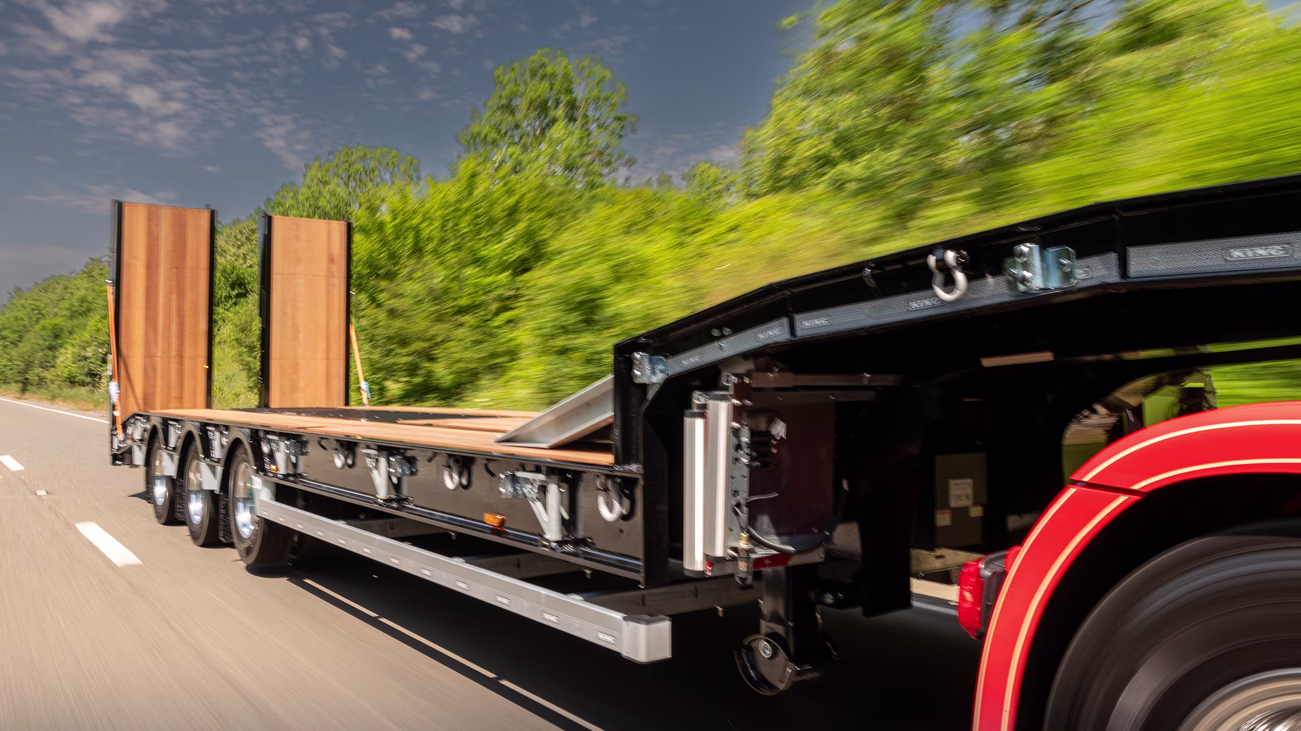 Flatbed trailer loaded with tall wooden beams on a highway, trees blurred by speed behind it.