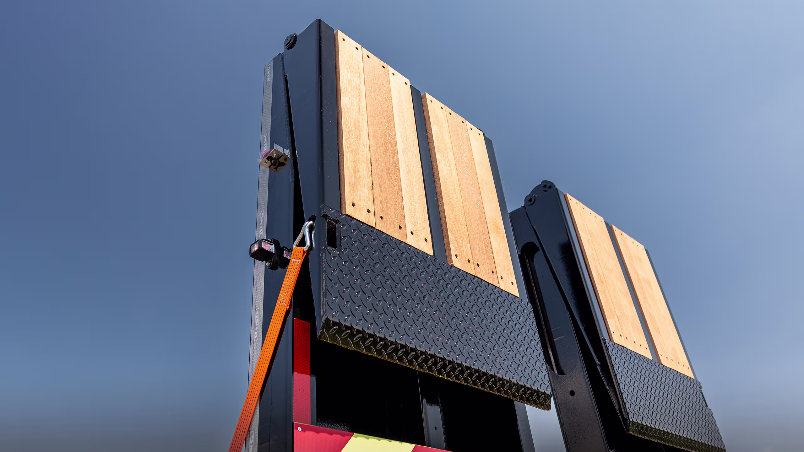 Close-up of a black steel truck trailer ramp with wooden planks, diamond-plate metal, and an orange strap against a clear blue sky