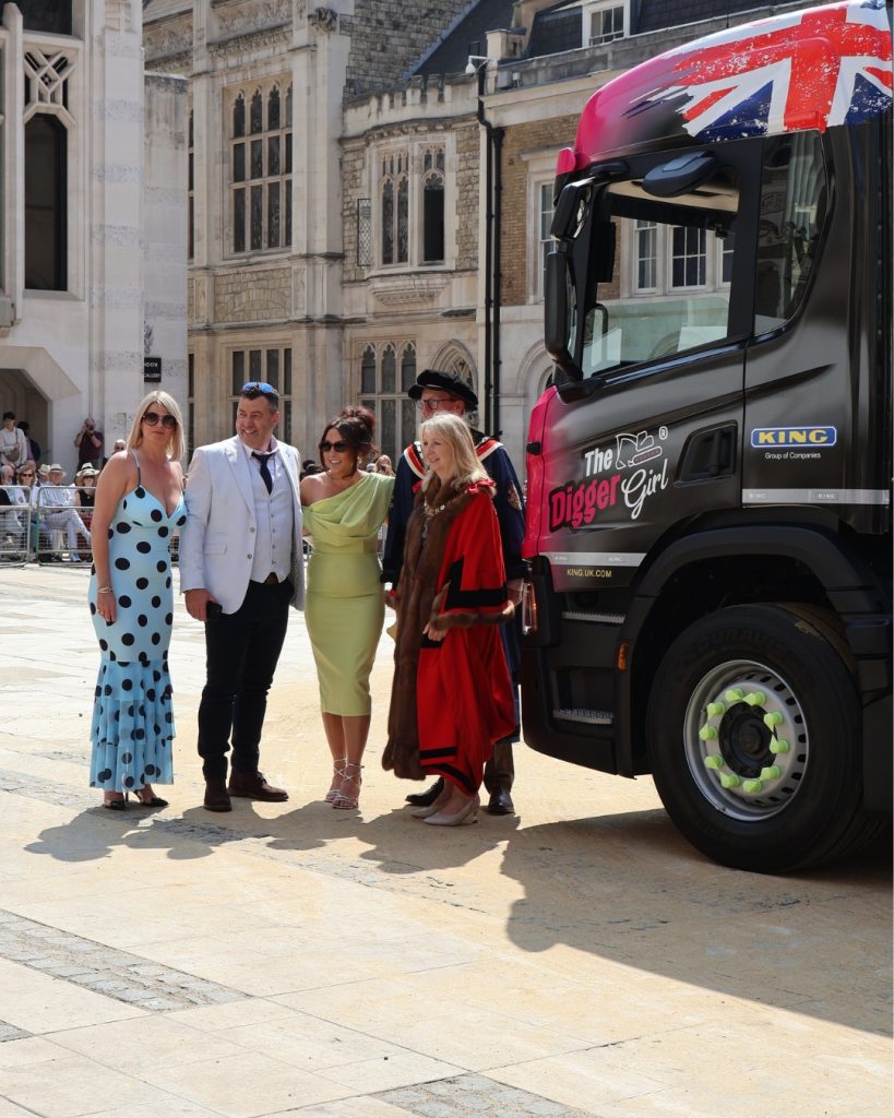Five adults in colorful outfits pose together beside a large black truck with a Union Jack wrap, in a historic stone-paved street.