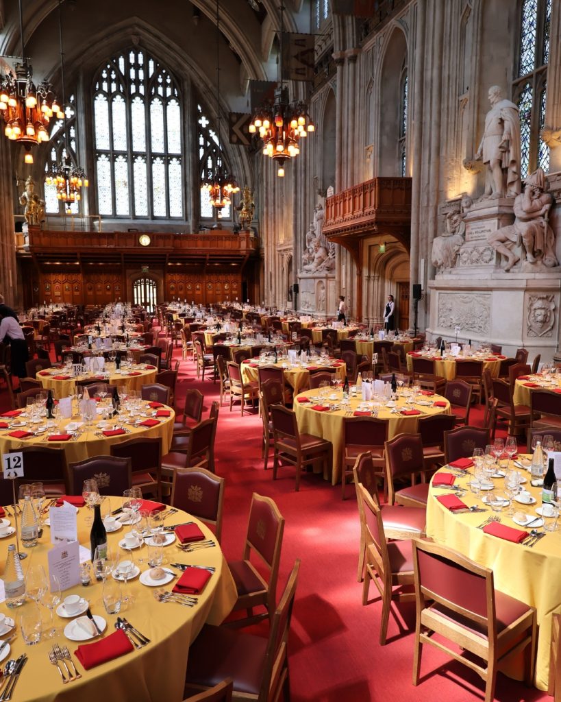 Banquet setup inside a grand cathedral-like hall with round tables covered in yellow cloths and red chairs under ornate chandeliers and statues/arches in the background, ready for a formal event.