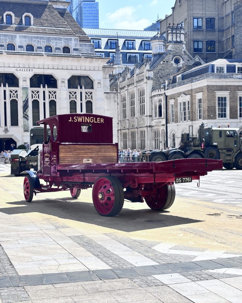 Vintage red flatbed truck with a closed cab labeled 'J. SWINGLER' parked in a city square near a historic gallery building; a crowd and modern vehicles appear in the background.