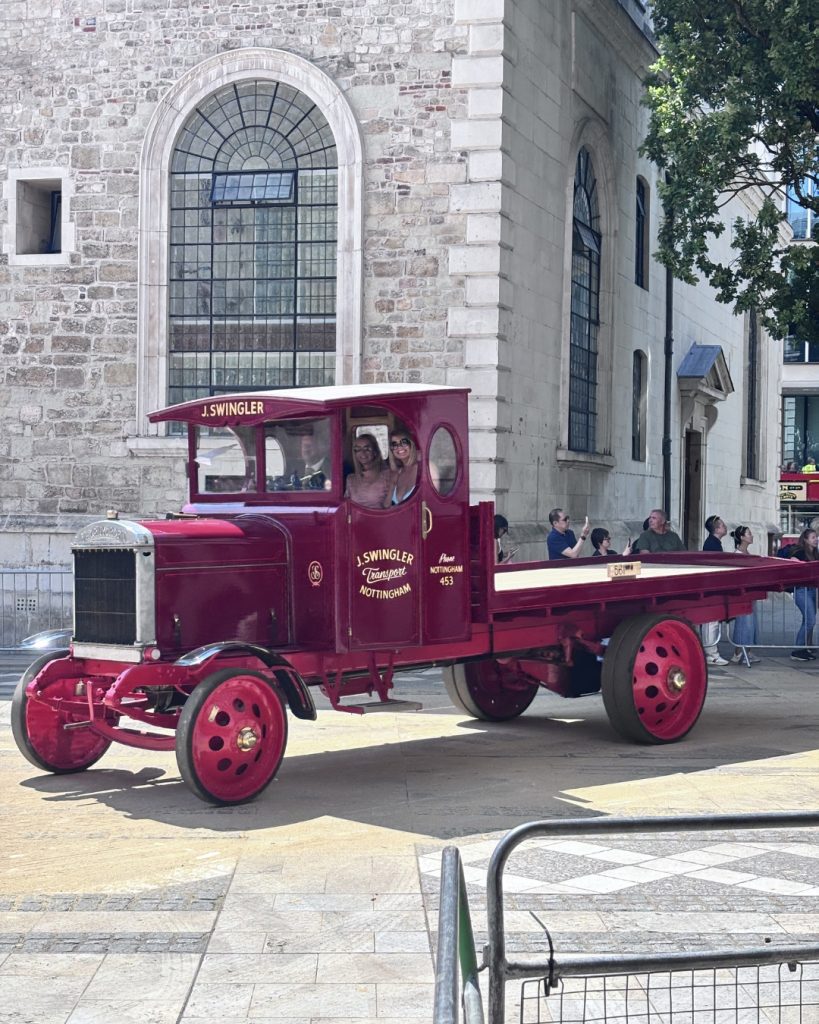 Vintage red transport wagon with an open cab and large spoked wheels, two women inside, parked along a stone building.