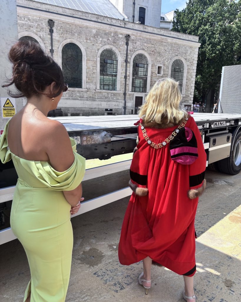 Two women in formal attire stand by a flatbed truck in front of a stone building with arched windows.