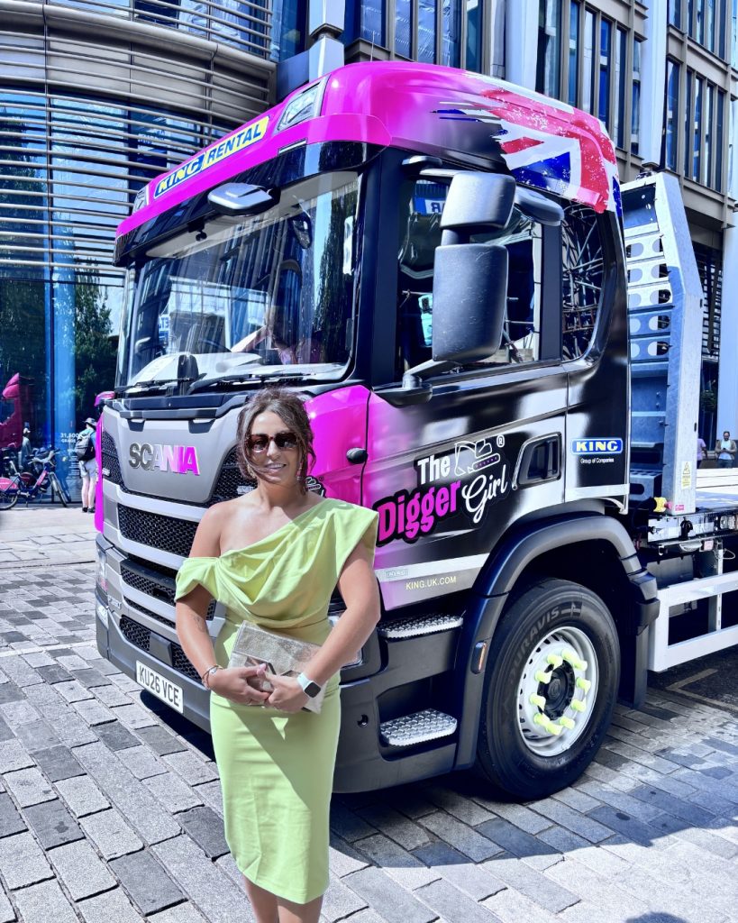 Woman in a lime-green one-shoulder dress posing in front of a bright pink Scania truck with 'The Digger Girl' branding in an urban plaza.