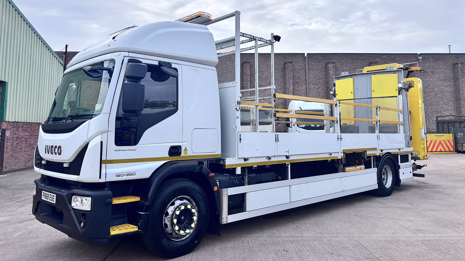 White Iveco Eurocargo truck with a long flatbed and yellow guard rails, parked in an industrial yard.