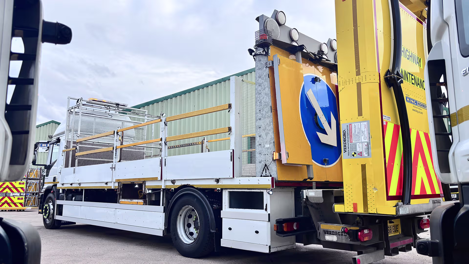 Large yellow highway maintenance truck with reflective chevrons and safety equipment, parked on a concrete lot in front of a green warehouse clamp and trailer gear in the background.