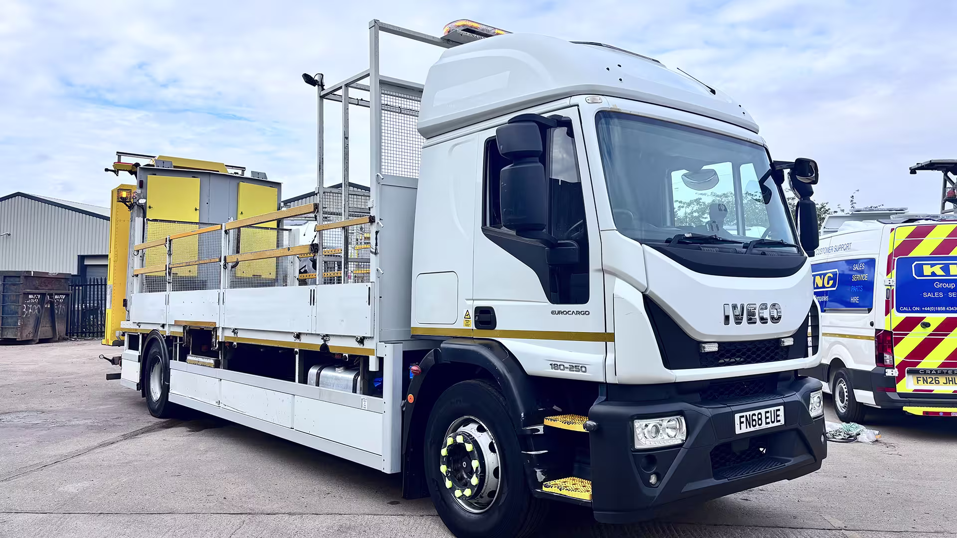 White Iveco Eurocargo flatbed truck in an industrial yard, with yellow machinery and metal fencing in the background.
