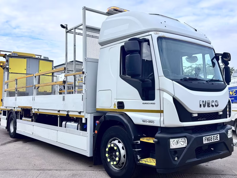 White Iveco Eurocargo flatbed truck in an industrial yard, with yellow machinery and metal fencing in the background.